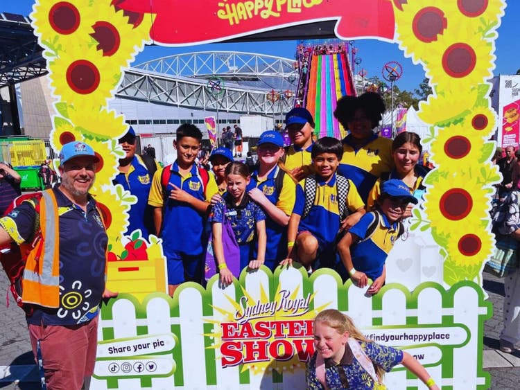 A group of smiling children stands in front of a sign that reads