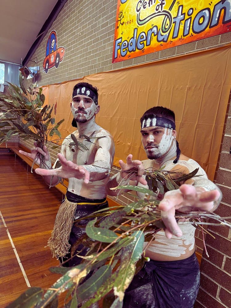 Two men in traditional attire hold large green leaves, standing together in a natural setting.